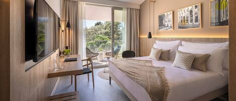 A modern hotel room with a large bed, a desk, and a balcony with a view of greenery.