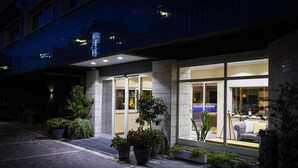 Una entrada de hotel moderna con fachada de vidrio, plantas en macetas y un interior bien iluminado visible a través de las ventanas.