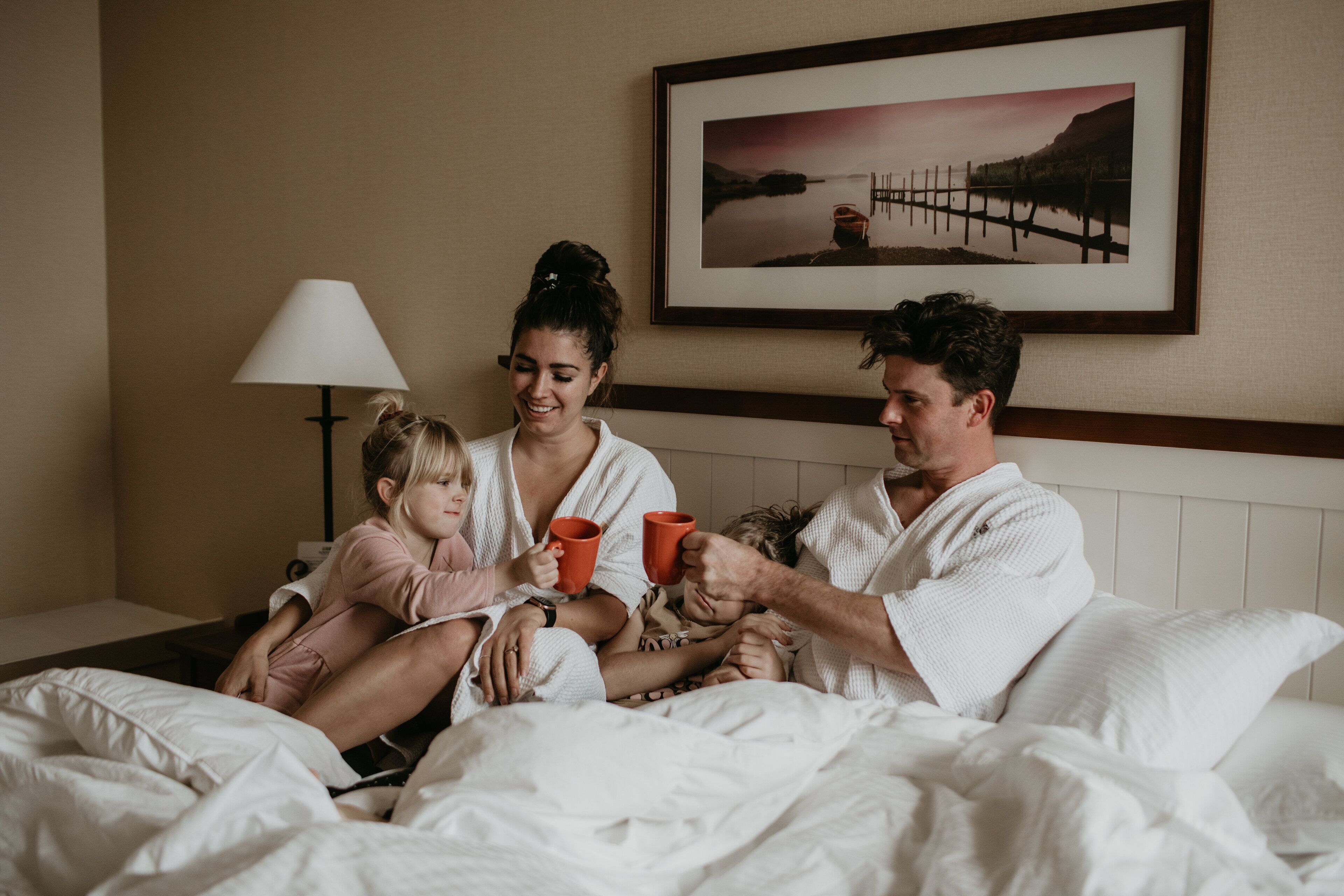 Una familia de cuatro en una habitación de hotel con una cama, una mesita de noche, una lámpara y un cuadro en la pared.