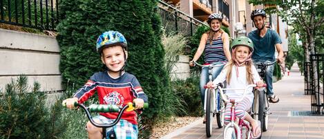 A family riding bicycles on a paved sidewalk next to a building with balconies.