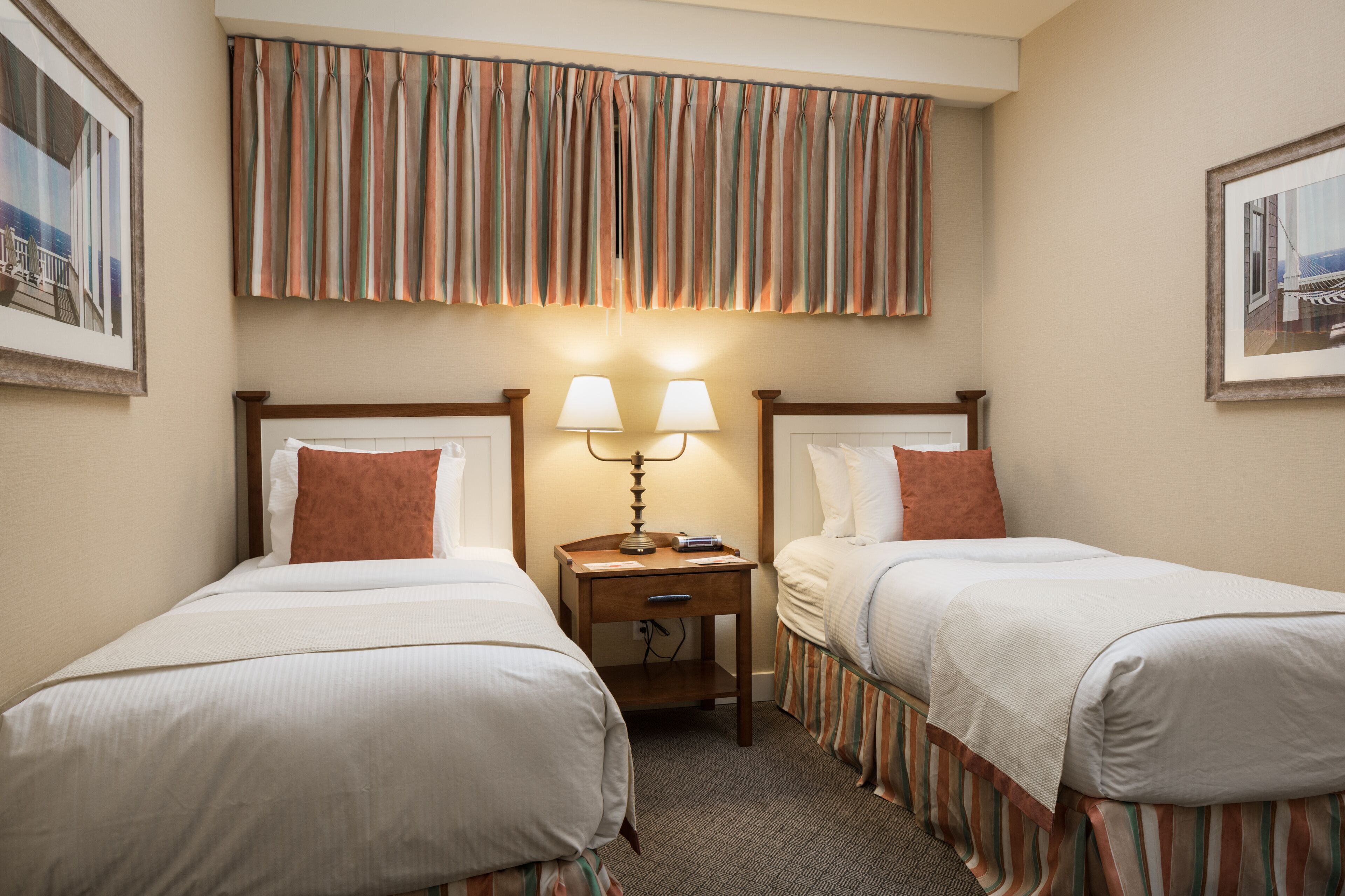A hotel room with two single beds, a nightstand with two lamps, and striped curtains.