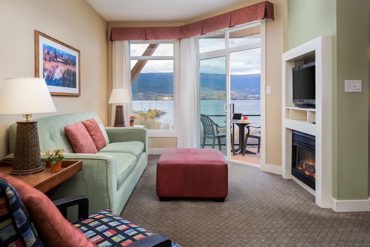 A hotel room with a green sofa, a red ottoman, a fireplace, a TV, and a view of a lake through large windows.