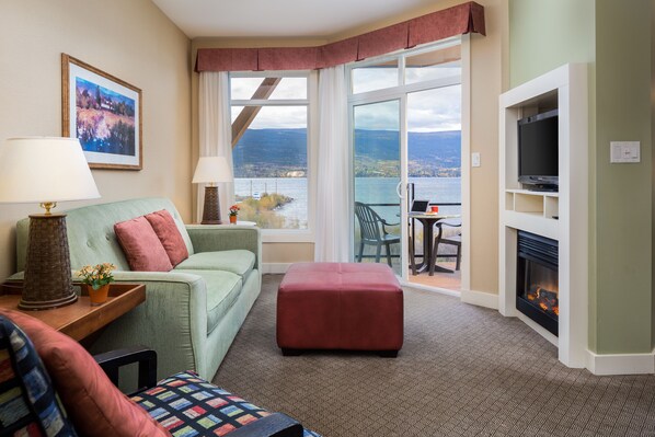 A hotel room with a green sofa, a red ottoman, a fireplace, a TV, and a view of a lake through large windows. - Summerland Waterfront Resort & Spa (Summerland)