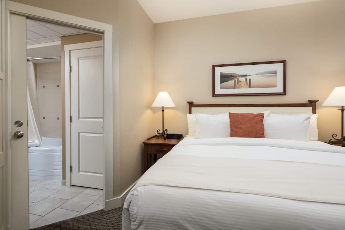 A hotel room with a large bed, two bedside lamps, a framed picture on the wall, and a bathroom visible through an open door.