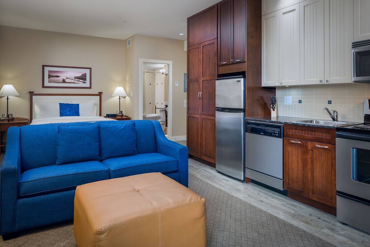 A hotel room with a blue sofa, an orange ottoman, a bed with white linens, a wooden cabinet, a stainless steel refrigerator, and a microwave.