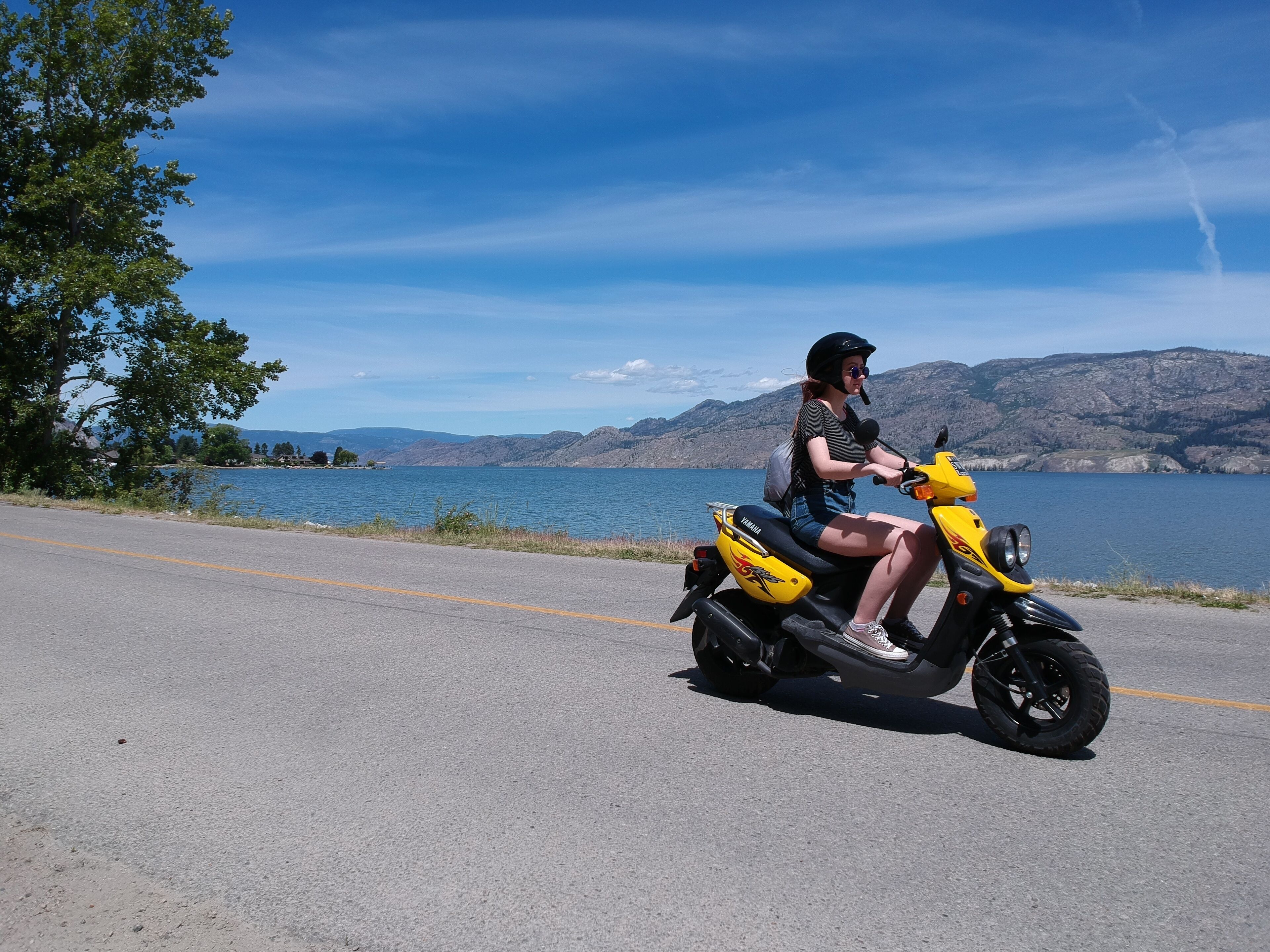 A person riding a yellow scooter on a road with a lake and mountains in the background.