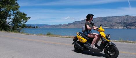 A person riding a yellow scooter on a road with a lake and mountains in the background.