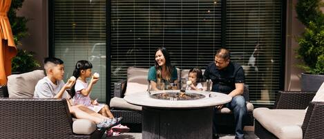 Een gezin van vijf personen zit rond een ronde tafel met een vuurkorf in het midden, op een terras met rieten meubilair en uitzicht op een gebouw door grote ramen.
