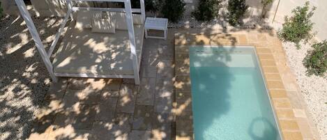 En swimmingpool med ett kaklat poolområde, en pergola med ett bambutak och små buskar längs poolens kant.