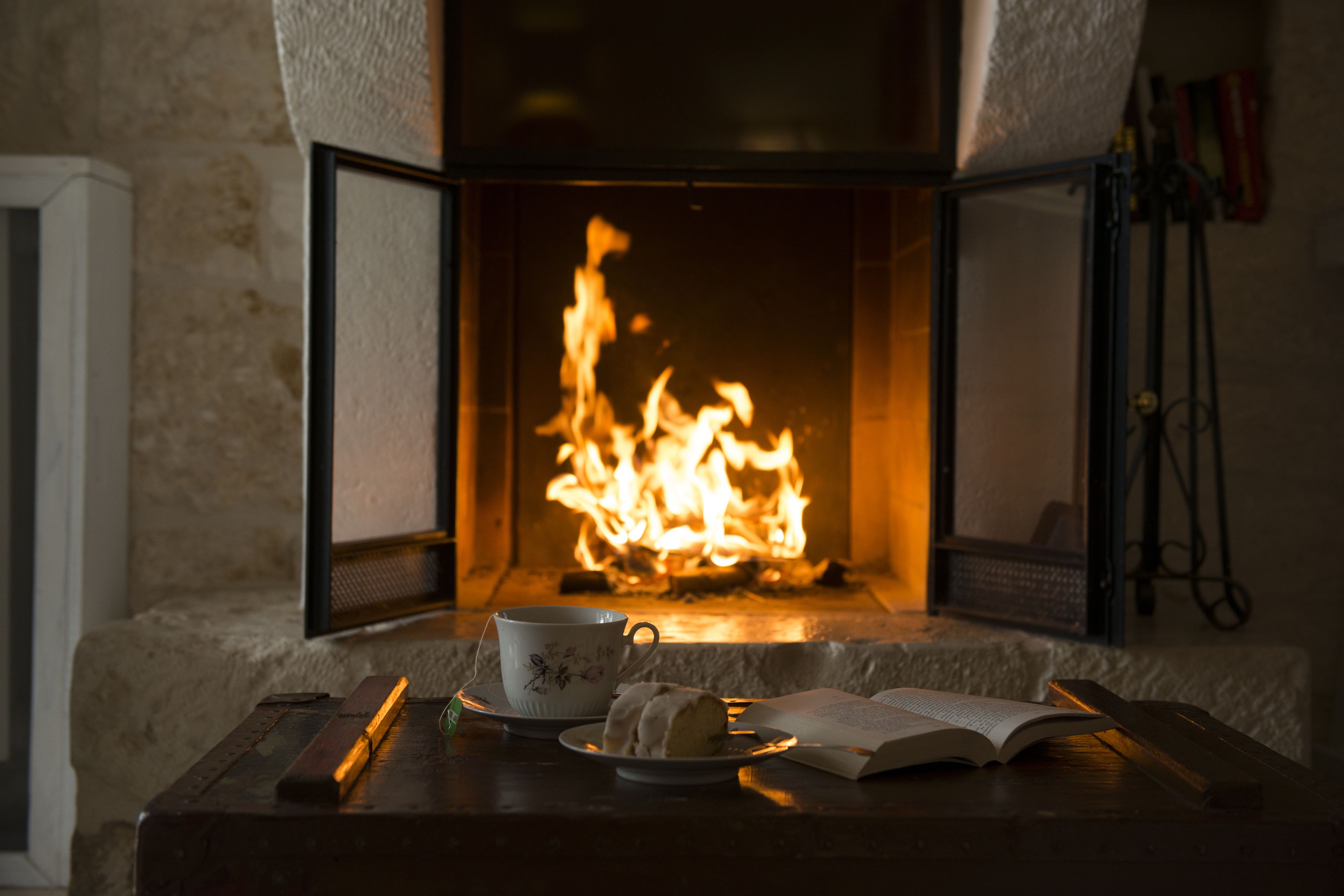 Una habitació acollidora amb una llar de foc, una tassa de te, un llibre i un plat amb un pastís damunt d'una taula de fusta.