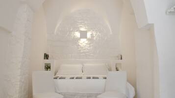 Una habitación blanca con una pared de piedra, una cama con ropa blanca y una mesita con una lámpara.