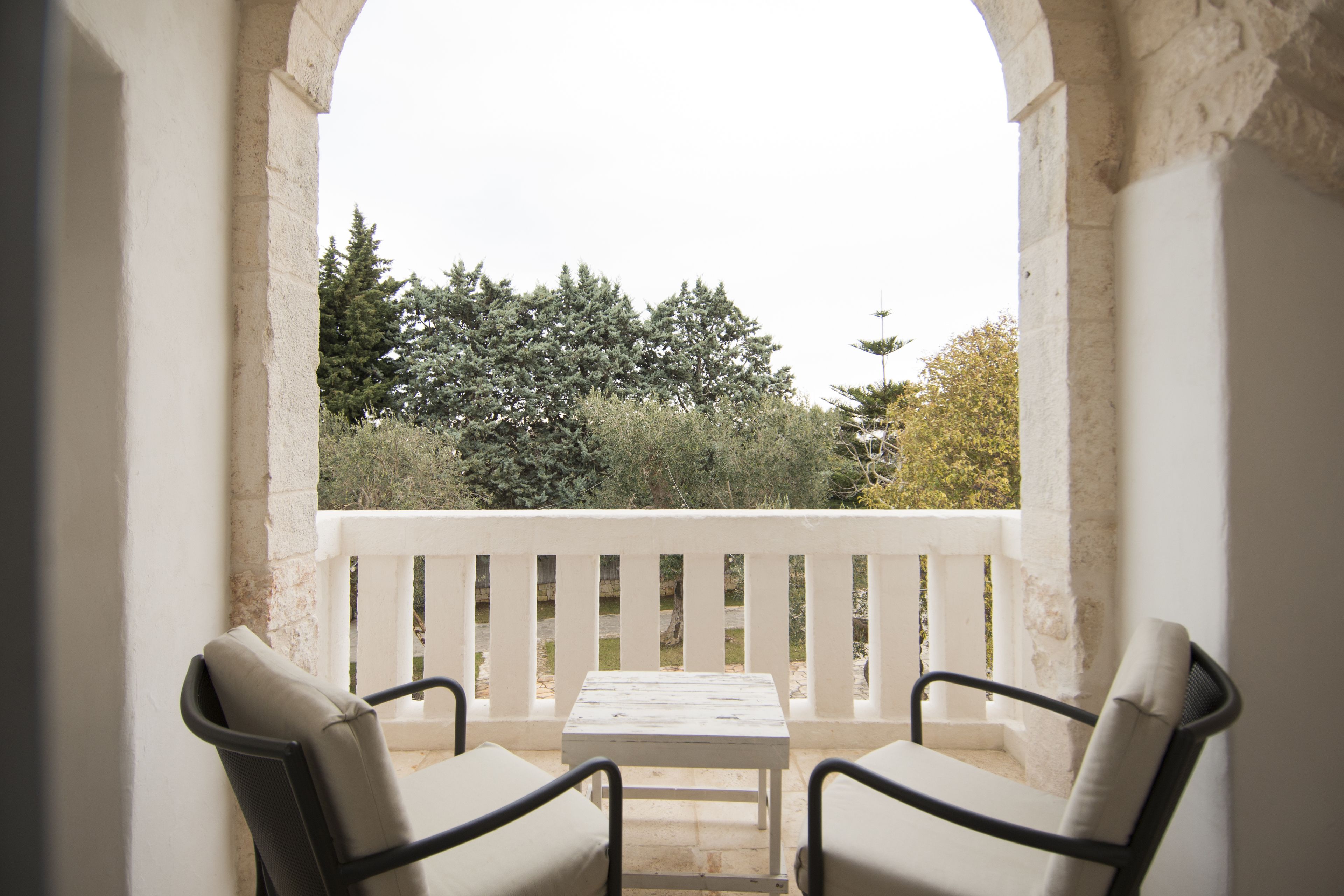 A balcony with two chairs and a table, overlooking a garden with trees and a white railing.