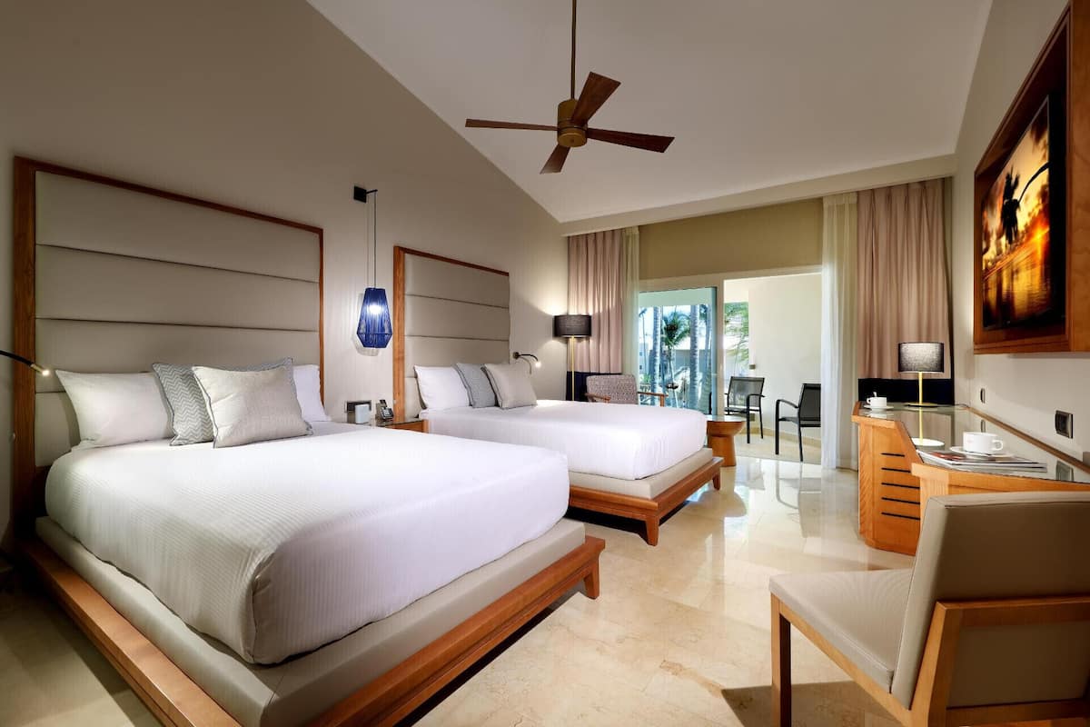 A hotel room with two beds, a ceiling fan, a desk with a chair, and a view of the outdoors.