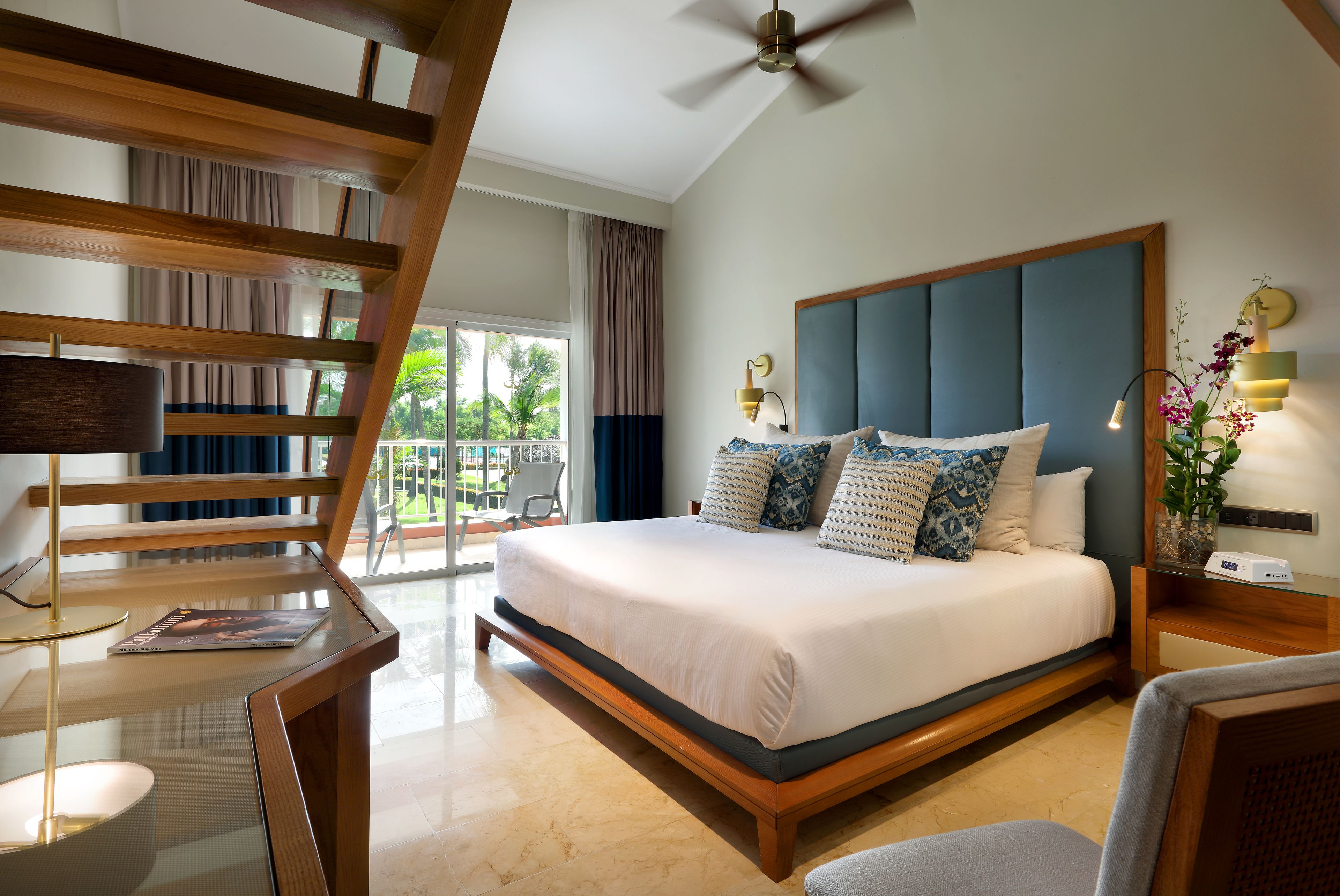 A modern hotel room with a large bed, a desk, and a balcony with a view of palm trees.