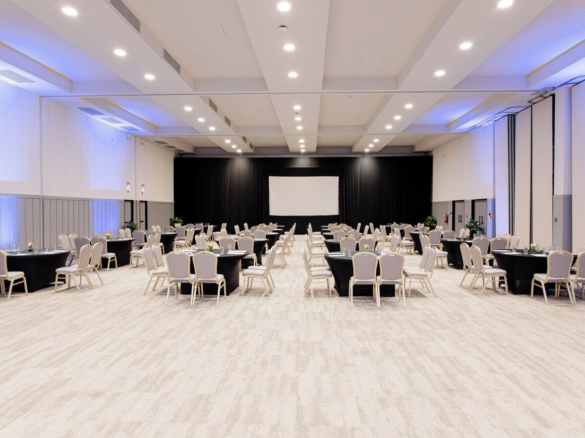 Un salón de conferencias con filas de mesas y sillas, una pantalla grande y un escenario.