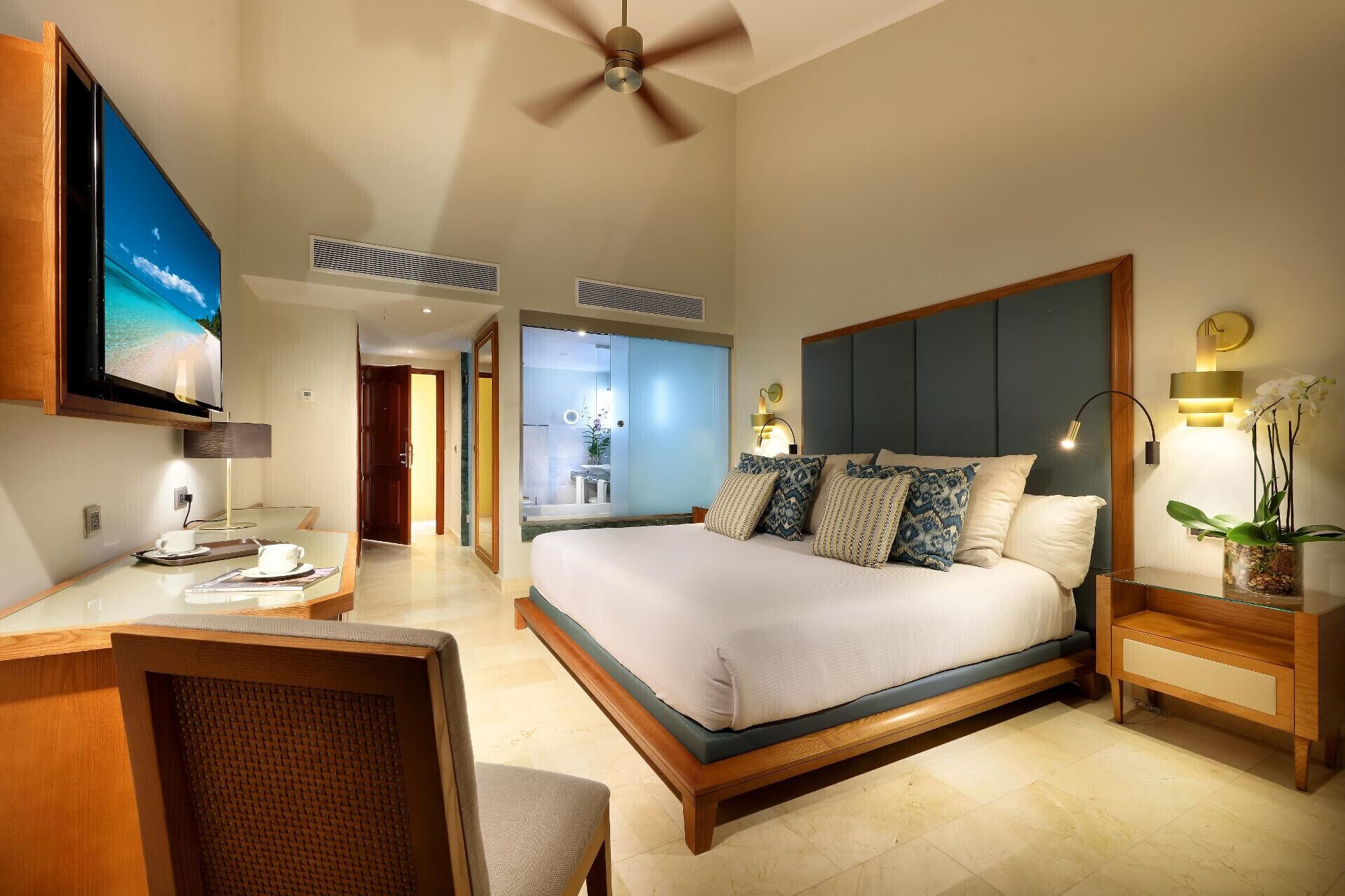 A hotel room with a bed, a desk, a chair, a TV, a ceiling fan, and a bathroom visible through an open door.