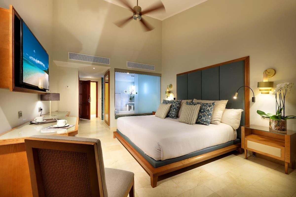 A hotel room with a bed, a desk, a chair, a TV, a ceiling fan, and a bathroom visible through an open door.