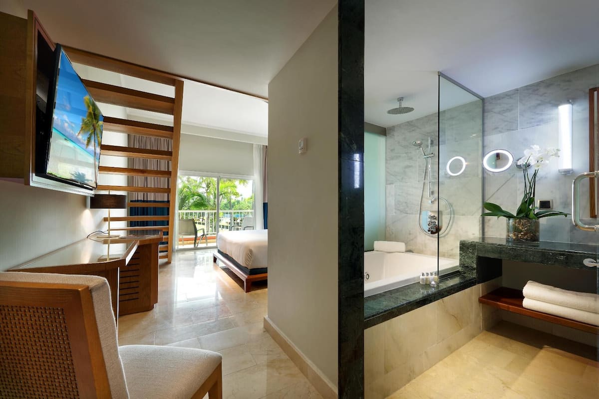 A modern hotel room with a balcony, a large bed, a TV, a bathroom with a bathtub, and a mirror.