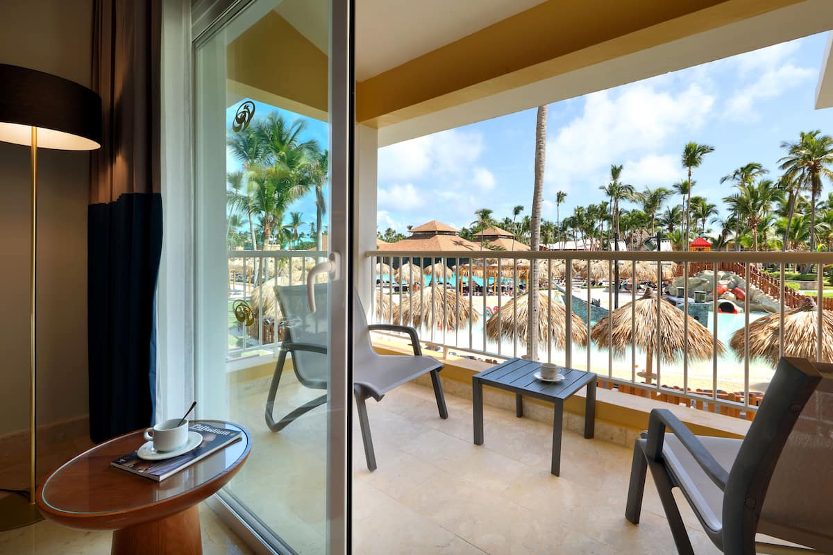 A balcony with a view of a resort pool, palm trees, and a thatched-roof structure.