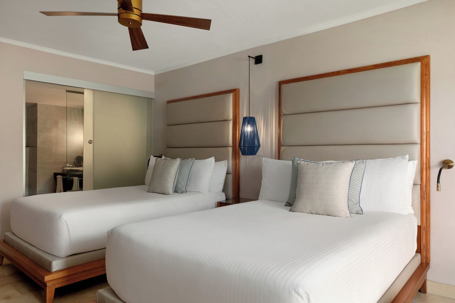Habitación de hotel con dos camas, ventilador de techo, lámpara colgante moderna y baño con espejo y toallero.
