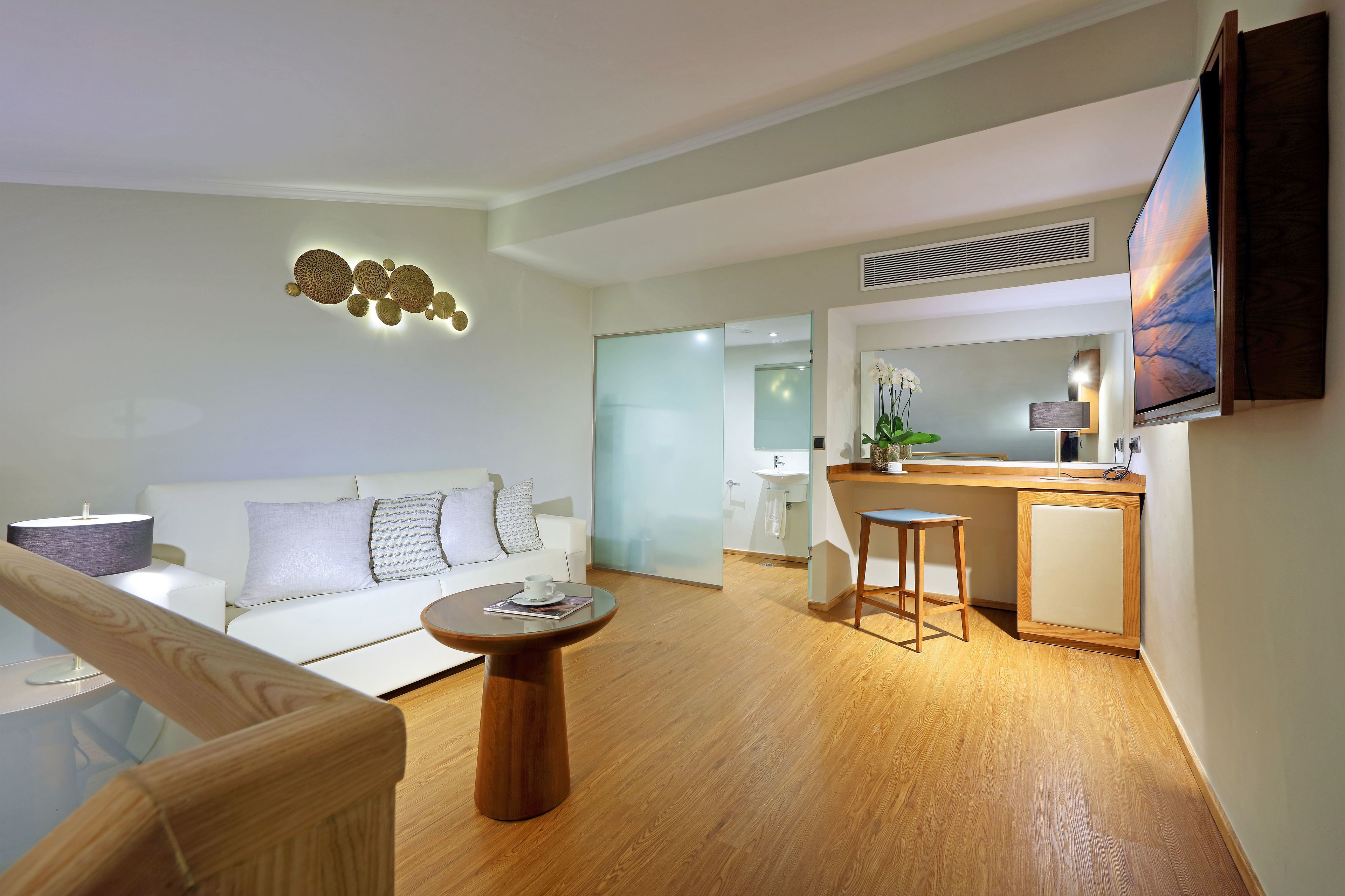 A modern living room with a white sofa, a round wooden coffee table, and a flat-screen TV mounted on the wall.
