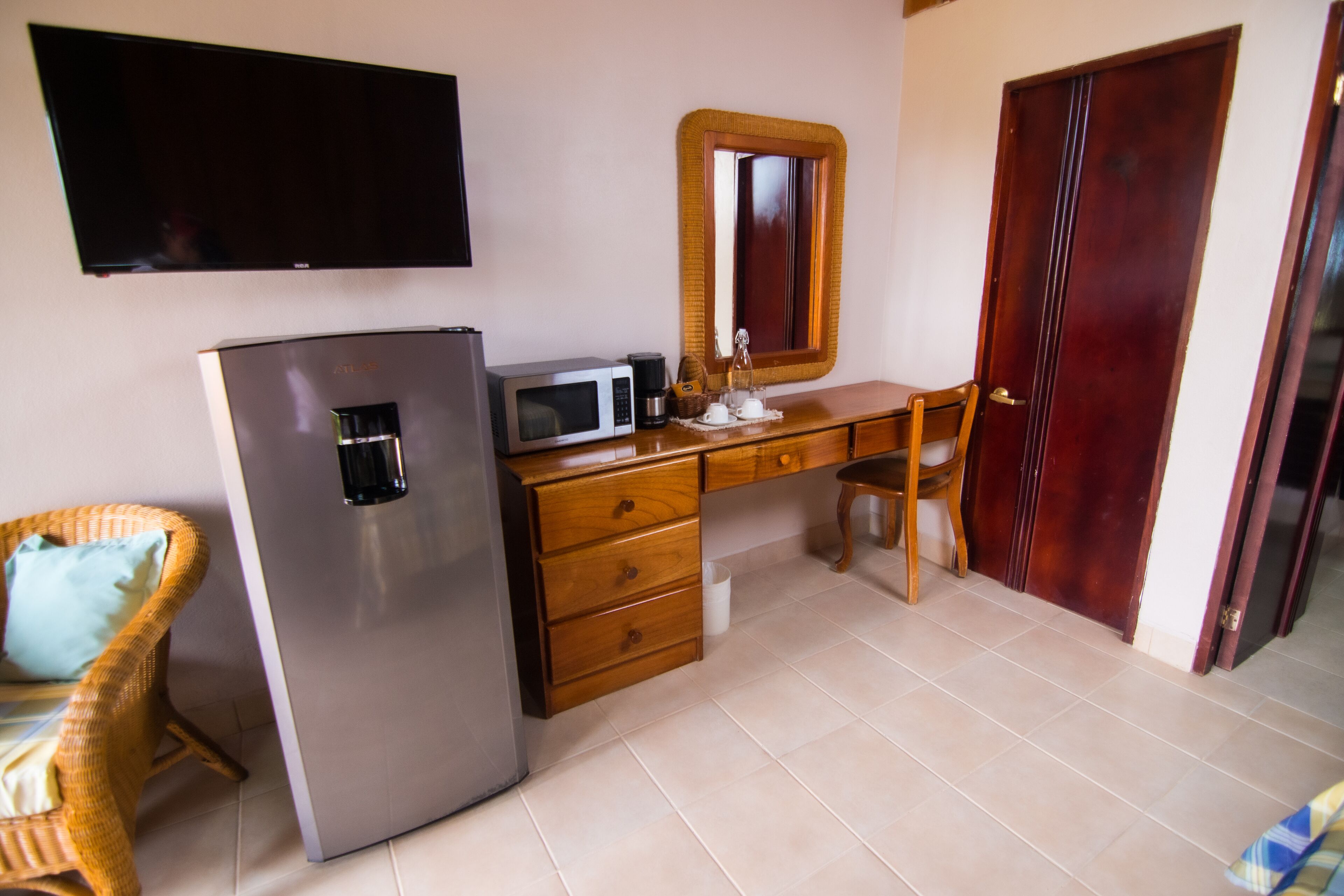 A compact kitchen with a refrigerator, microwave, wooden desk, wicker chair, and a television mounted on the wall.