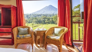 Une pièce avec des fauteuils en osier et une petite table, une grande fenêtre avec des rideaux rouges, et une vue sur une montagne.