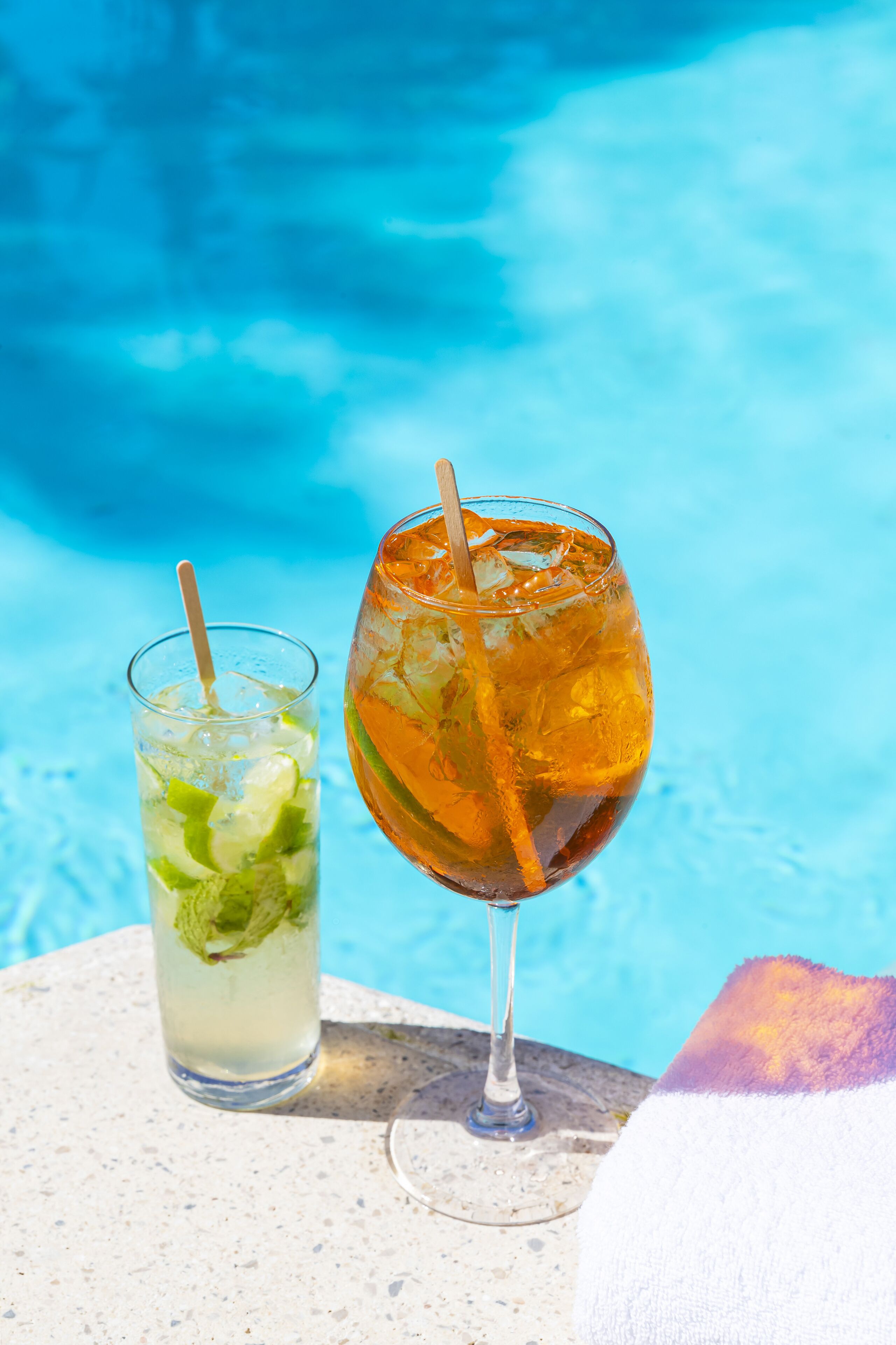 Dos bebidas junto a una piscina, una en un vaso alto y otra en una copa de vino, con una piscina azul cristalina al fondo.