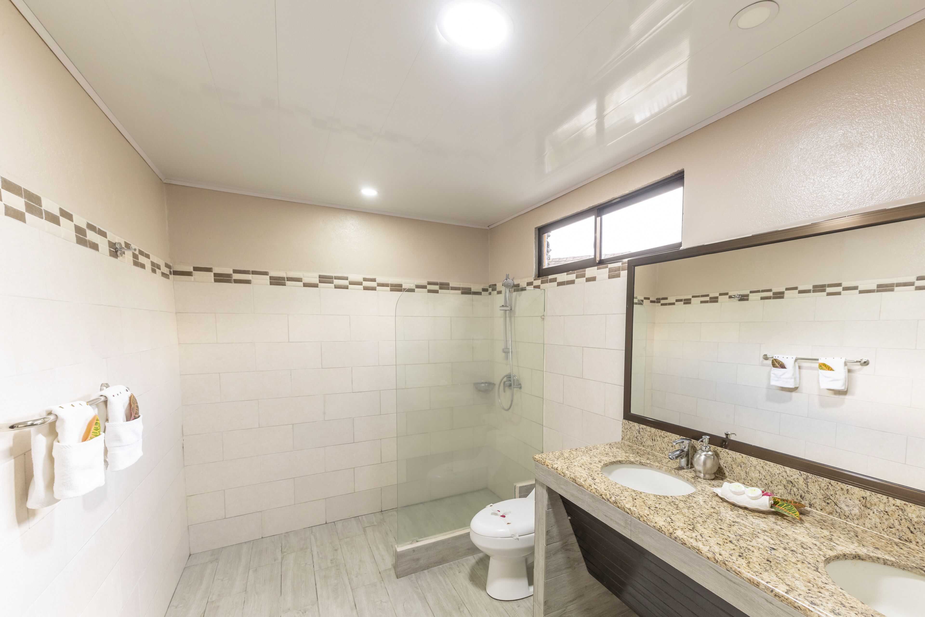 Un baño moderno con ducha envuelta en vidrio, inodoro blanco y encimera de granito con dos lavabos.