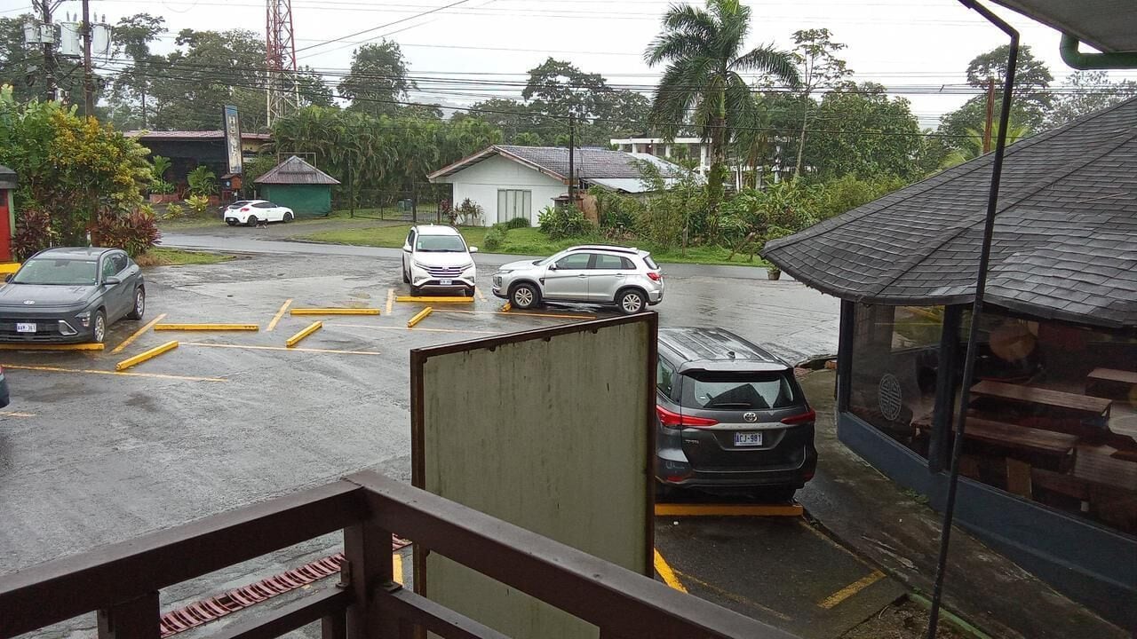 A parking area with several cars, a small building, and greenery in the background.