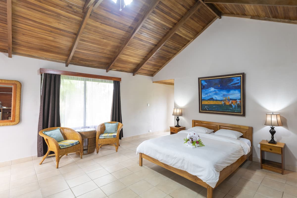 Een slaapkamer met een houten plafond, een bed met witte lakens, twee nachtkastjes met lampen, een rieten stoel, een klein tafeltje en een raam met gordijnen.