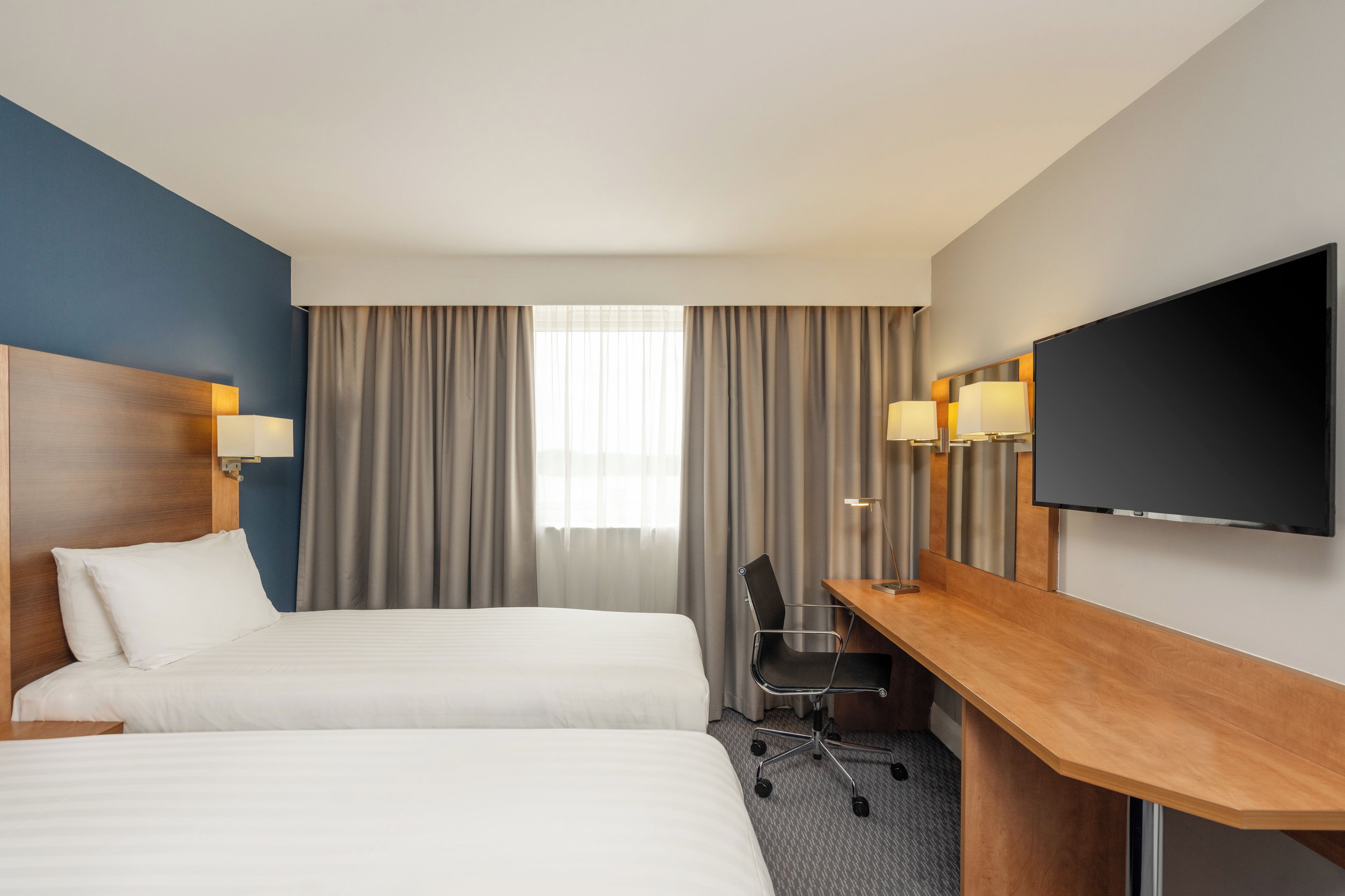 Quarto de hotel com duas camas, uma mesa, uma cadeira, televisão de tela plana e janela com cortinas.
