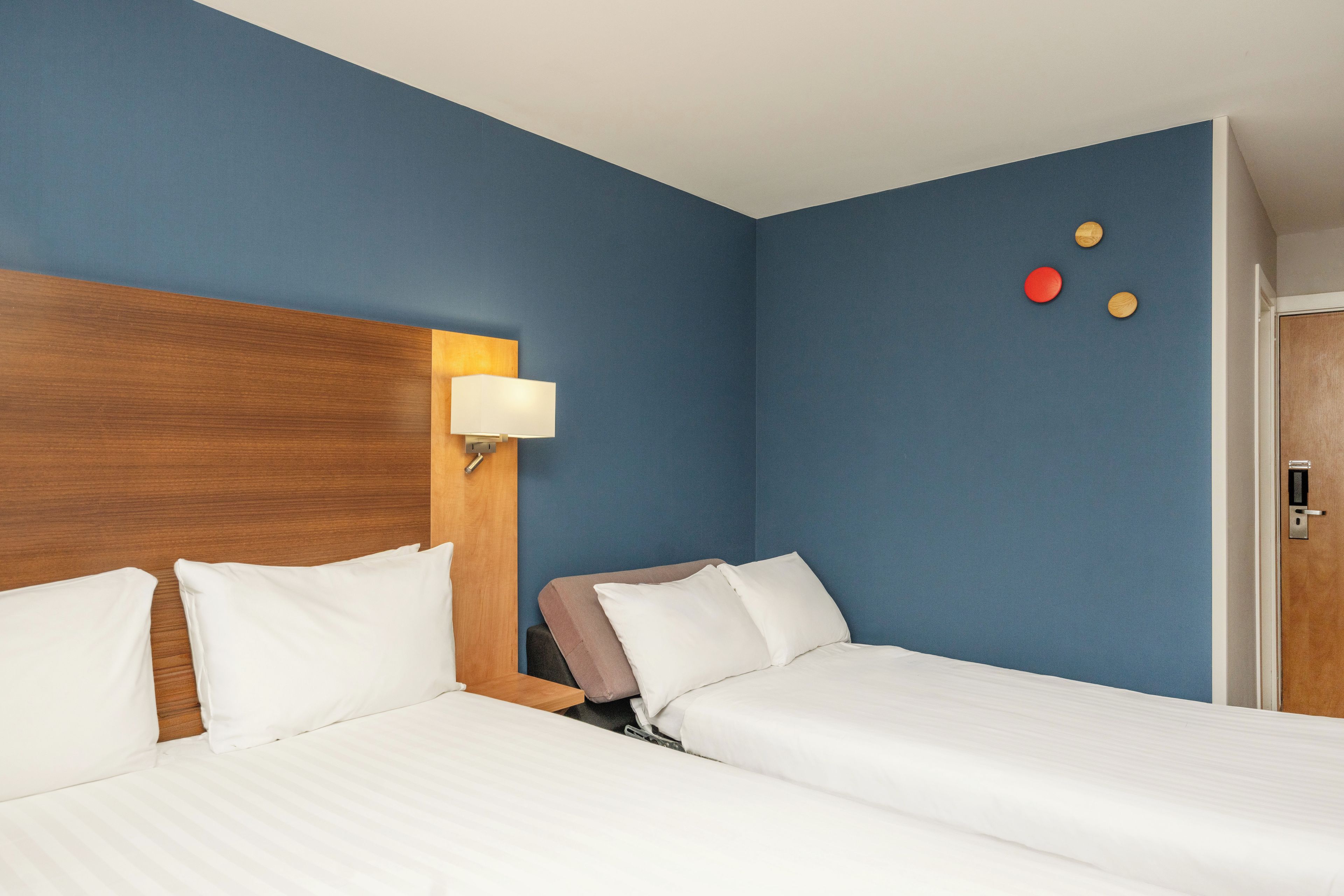 Habitación de hotel con dos camas, cabecero de madera, mesita de noche y lámpara de pared.