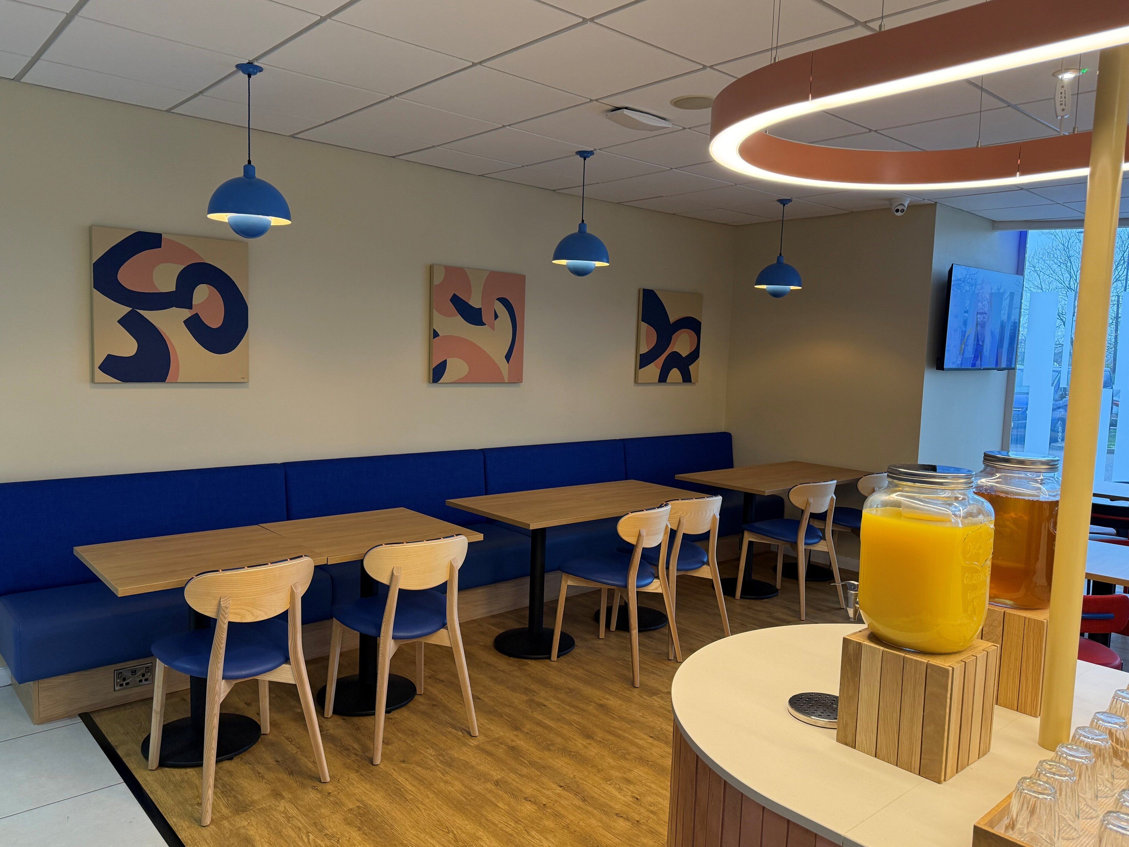 Un interno di un caffè moderno con sedute blu, tavoli in legno, opere d'arte astratte al muro e un angolo dedicato ai succhi di frutta.