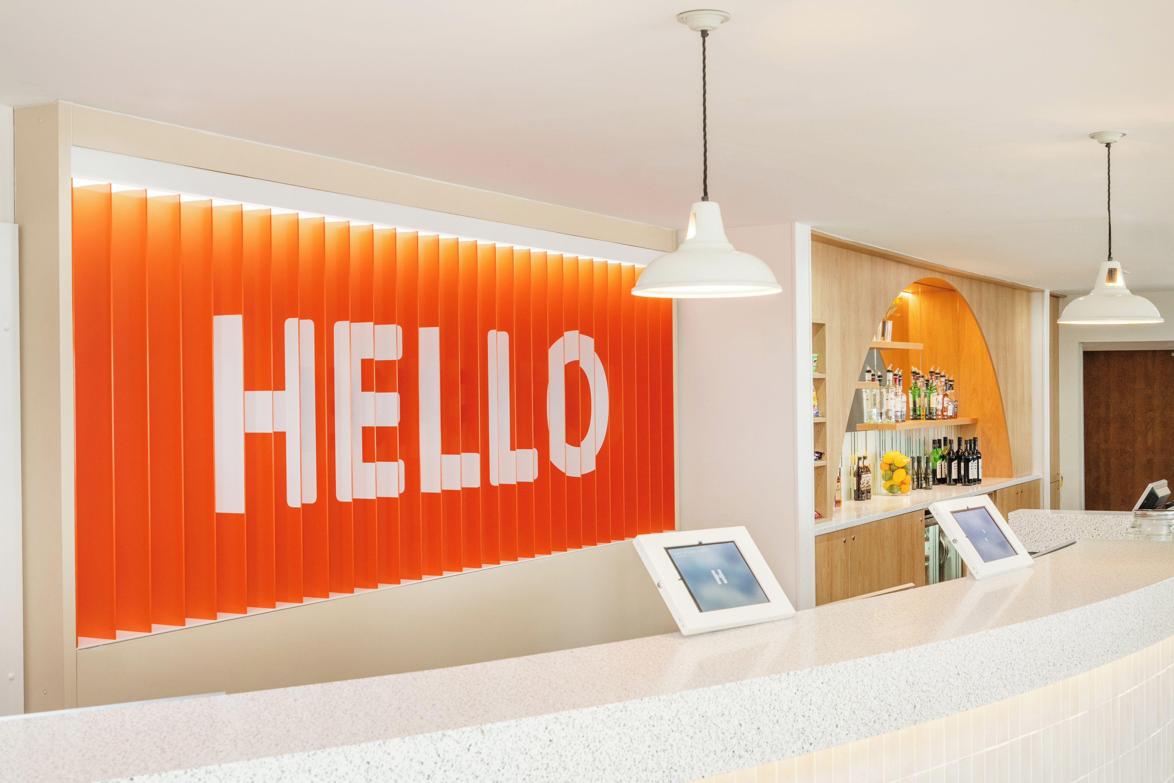 Zona de recepción con un cartel grande que dice 'HELLO', un mostrador con pantalla digital y una zona de bar con botellas.