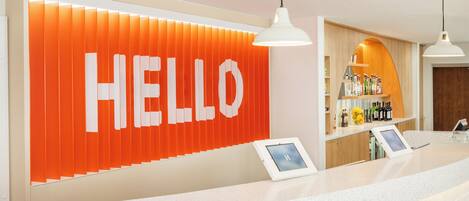 Zona de recepción con un cartel grande que dice 'HELLO', un mostrador con pantalla digital y una zona de bar con botellas.