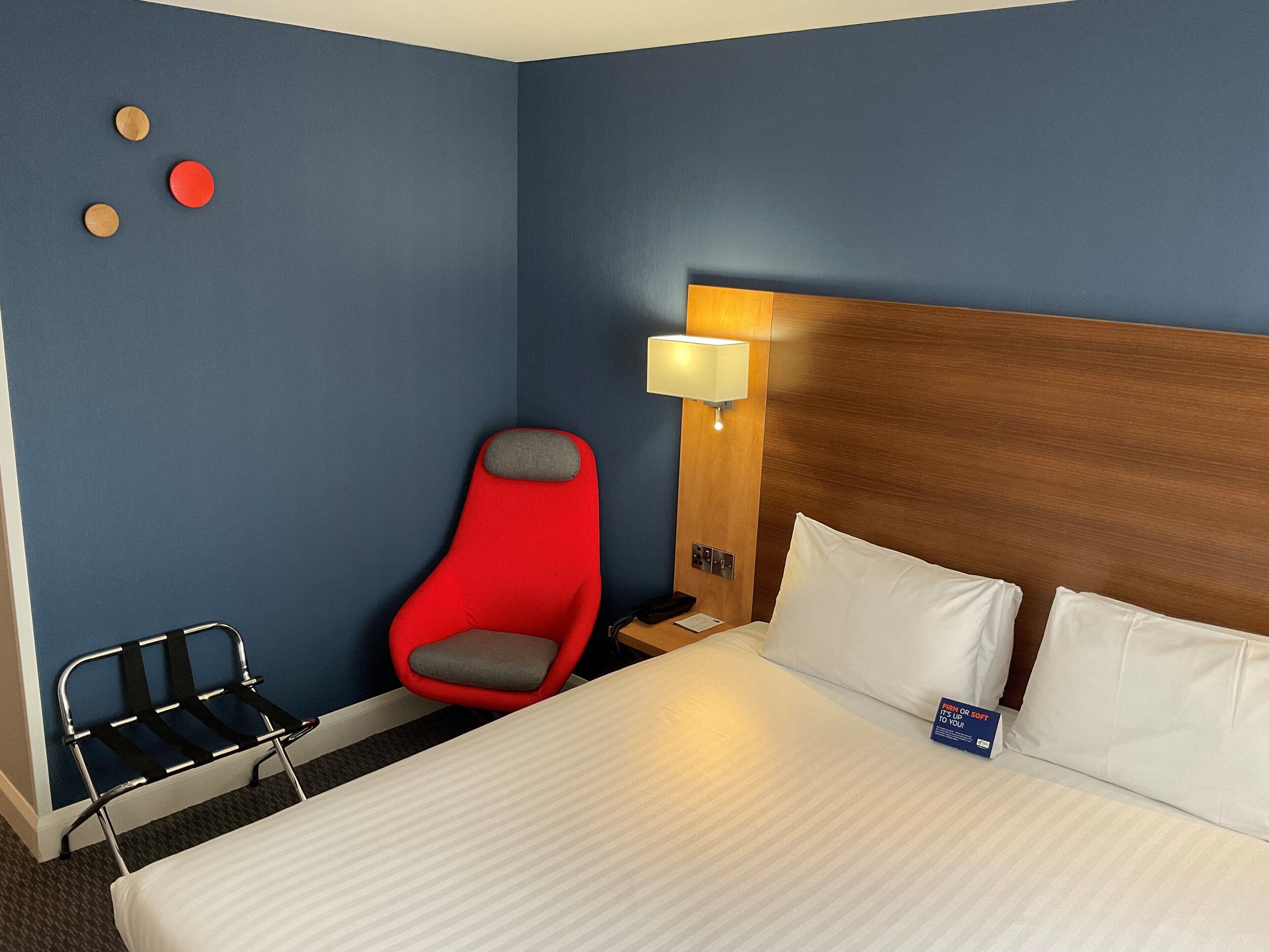 Habitación de hotel con una cama grande, una silla roja, una mesita de noche con lámpara y un reloj en la pared.