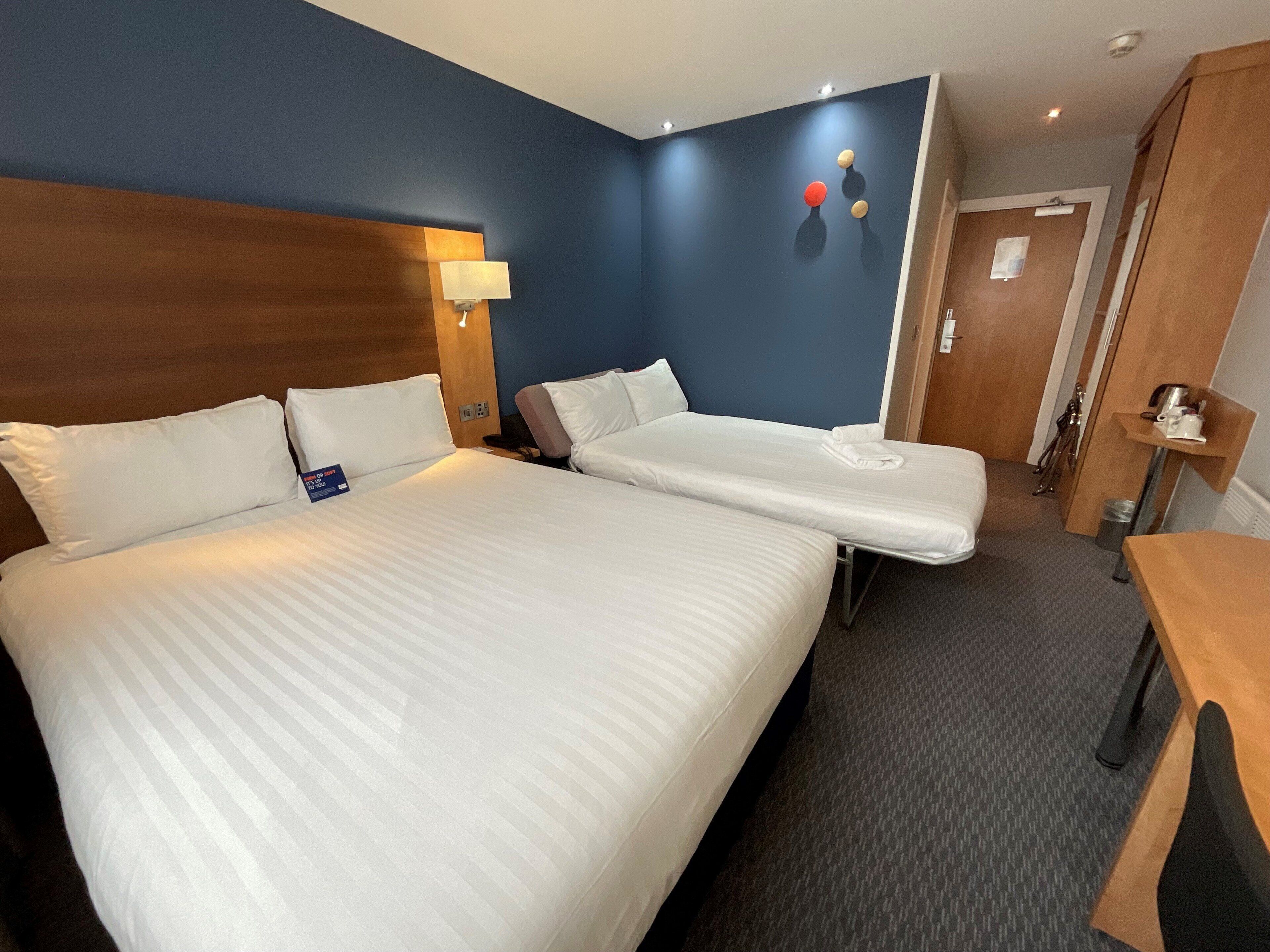A hotel room with two beds, a wooden headboard, a nightstand, a lamp, and a small table with a cup.