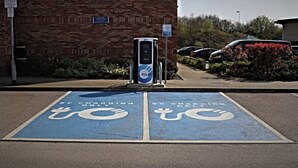 A parking area with EV charging stations, marked spaces, and a brick building in the background.