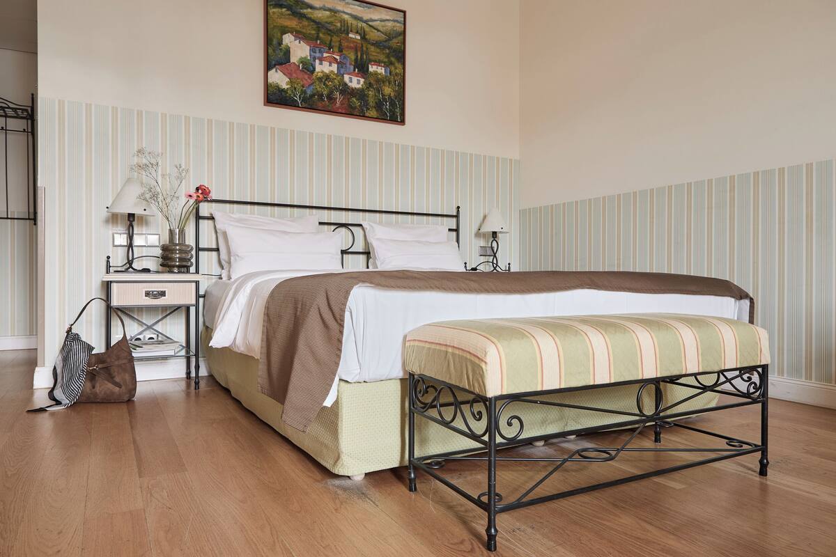 Un dormitorio con cama, mesitas de noche, lámpara, un cuadro en la pared y suelo de madera.