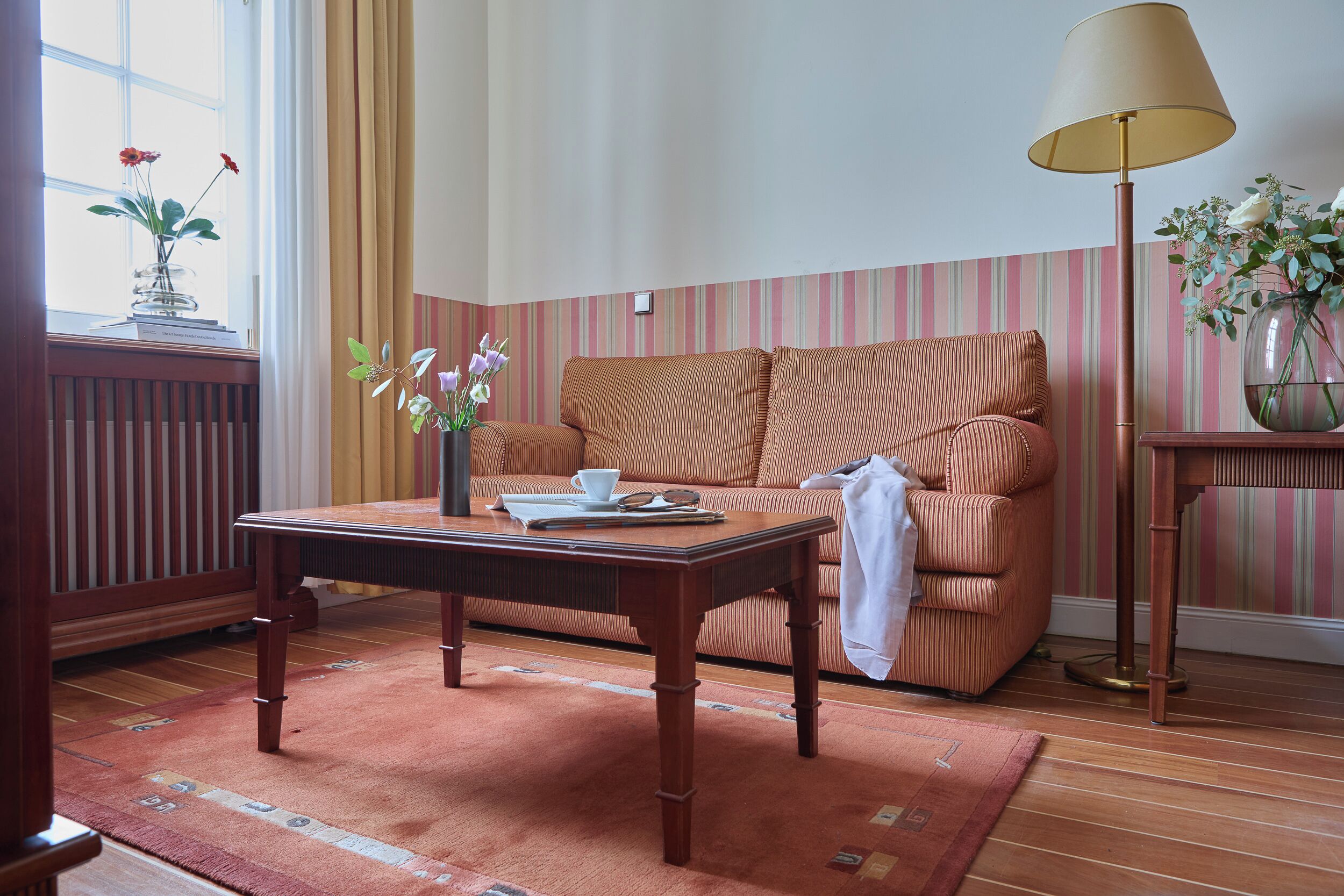 Una habitación con una mesa de madera, un sofá a rayas, una lámpara y un florero con flores.