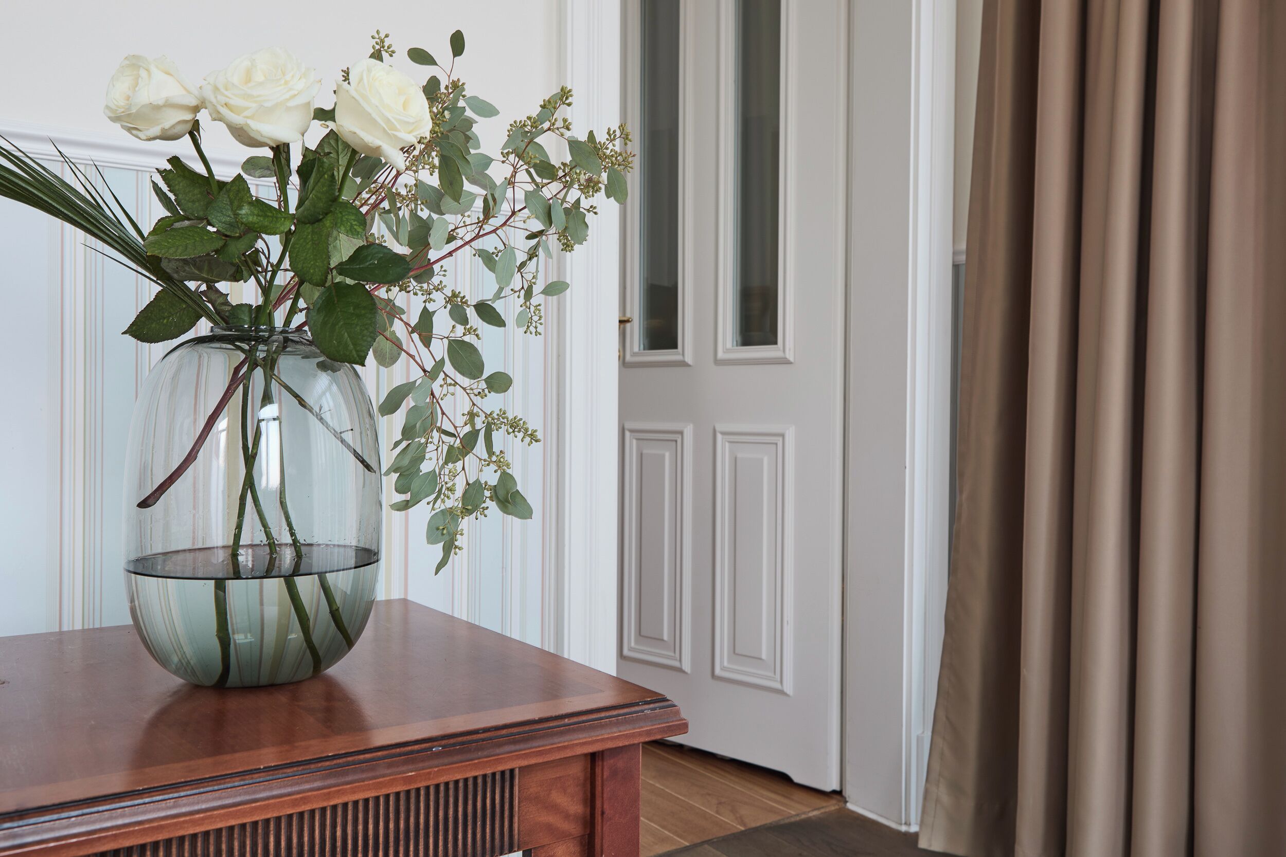 Vista despejada de una habitación con una mesa de madera, un jarrón de vidrio con flores blancas y una puerta con cortinas.