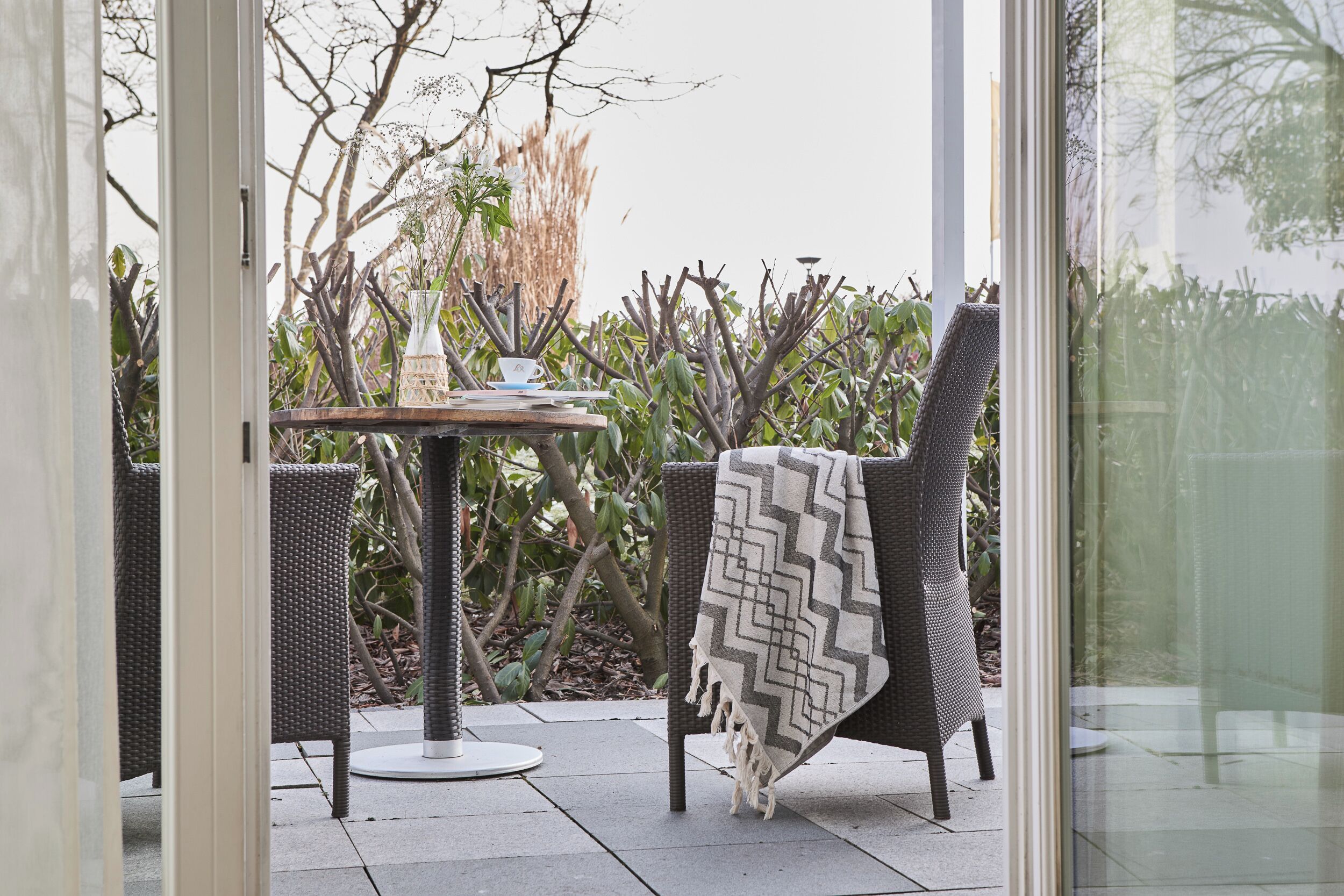 Eine Terrassenfläche mit Korbmöbeln, einem Tisch mit einer Vase und Tasse sowie einem Sessel mit dekorativer Tagesdecke.