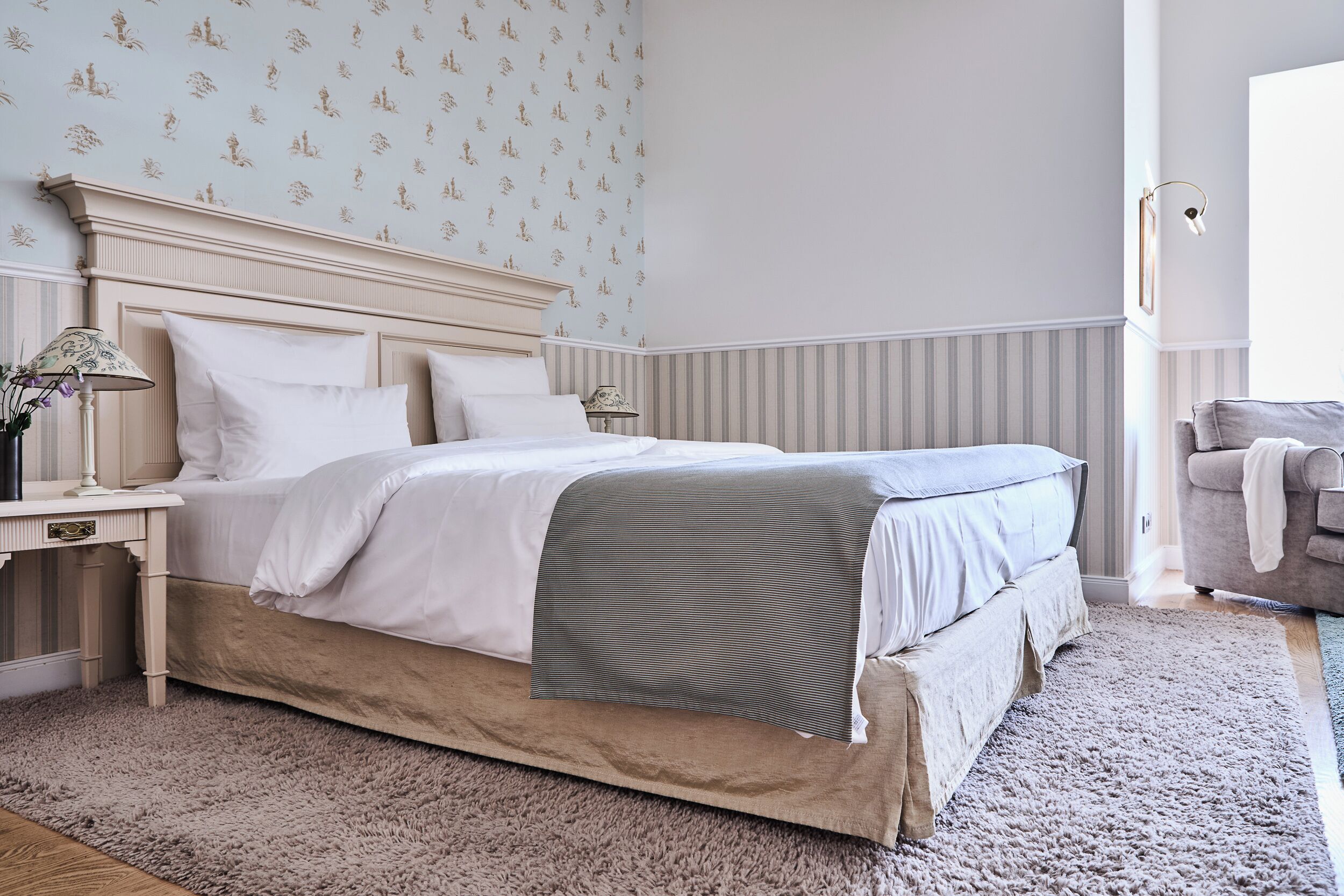 A bedroom with a bed, nightstands, a lamp, a chair, and a patterned wall.