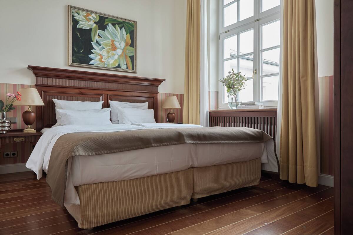 Una habitación de hotel con una cama grande, cabecero de madera, dos lámparas de noche, una ventana con cortinas y un cuadro con motivos florales en la pared.