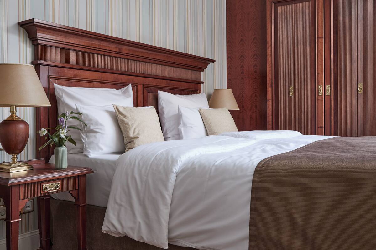 Habitación de hotel con cabecera de madera, cama con ropa blanca, mesita de noche con lámpara y un florero con flores.