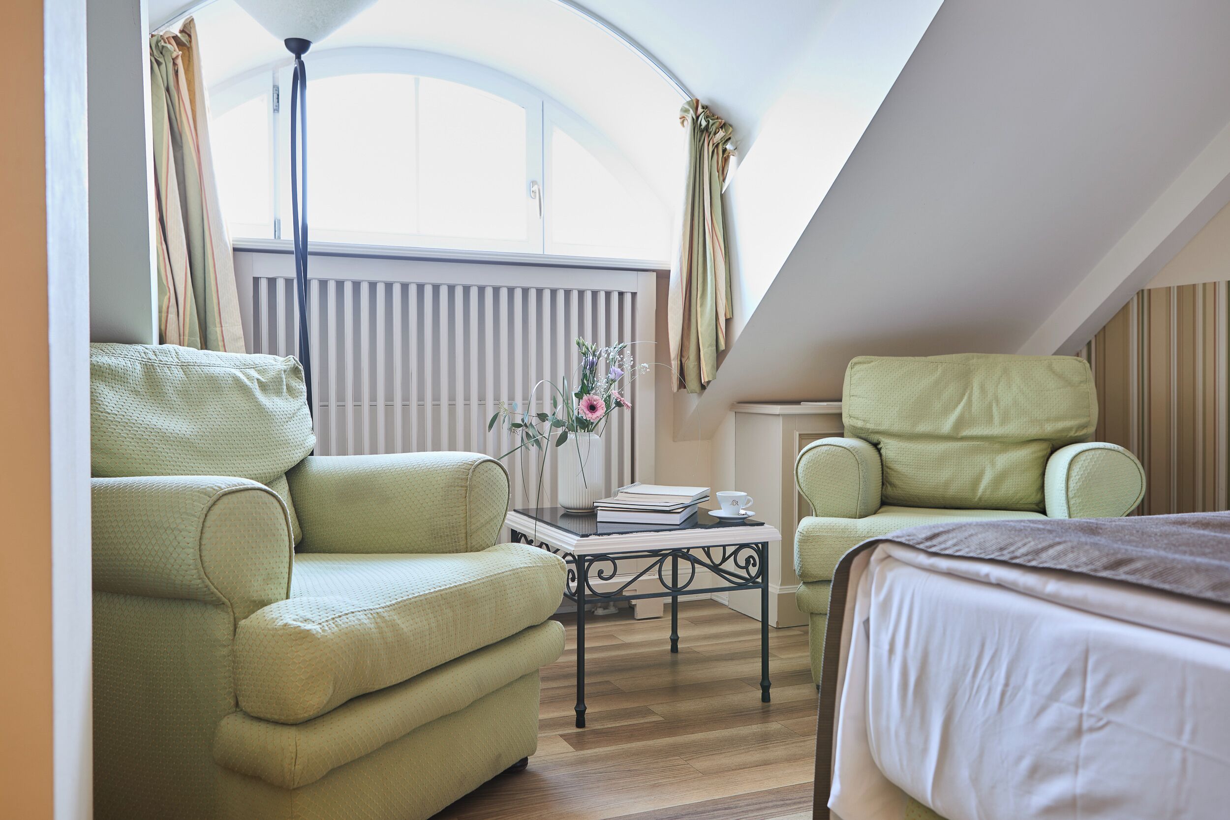 Ein Zimmer mit einem Bett, zwei grünen Sesseln, einem kleinen Tisch mit Blumenvase und einem Heizkörper.