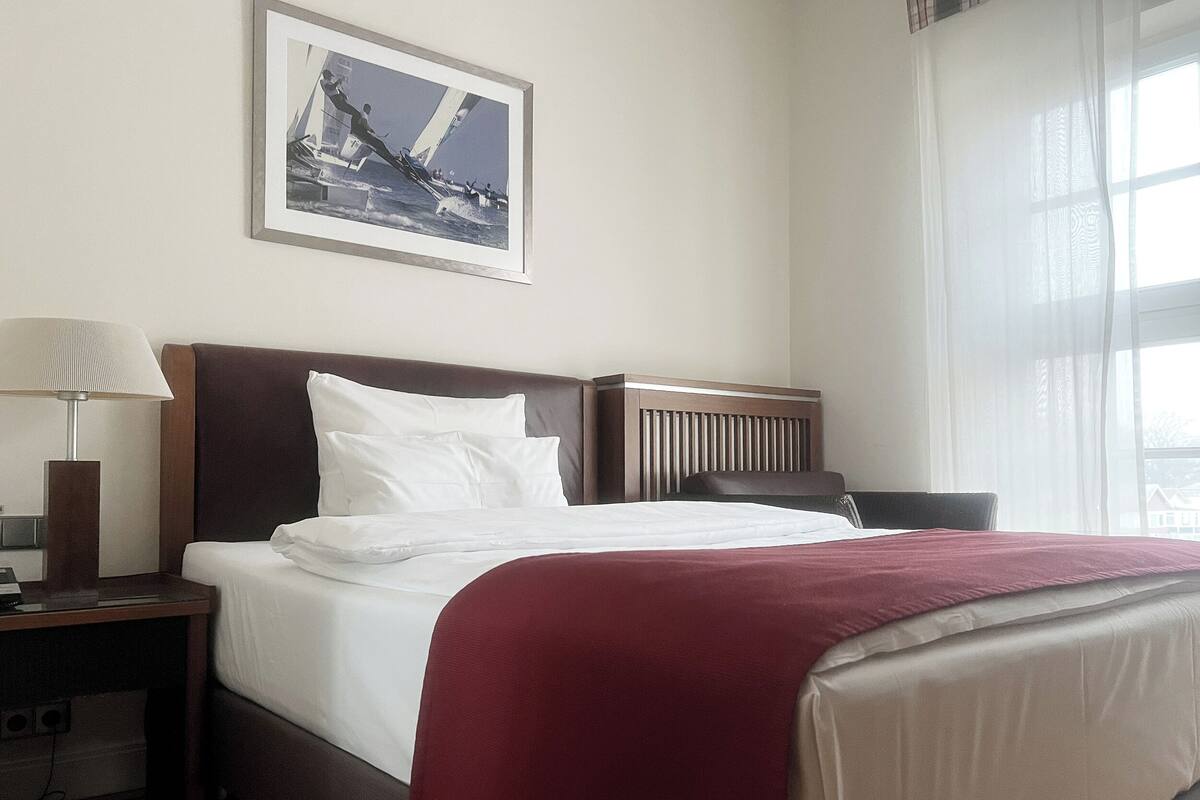 Un cuarto de hotel con una cama, una mesita de noche, una lámpara, una ventana con cortinas translúcidas y un cuadro en la pared.