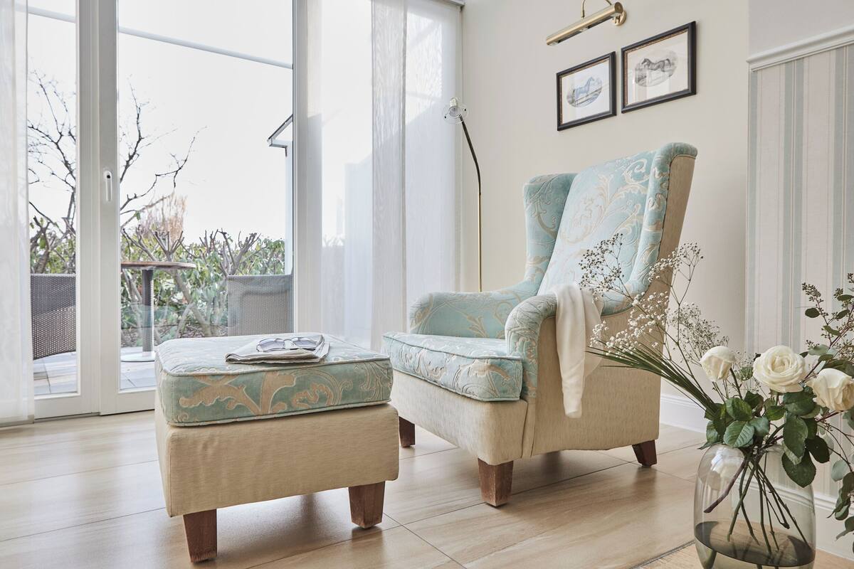 Una habitación con un ventanal grande, una silla, un reposapiés y un florero con flores.