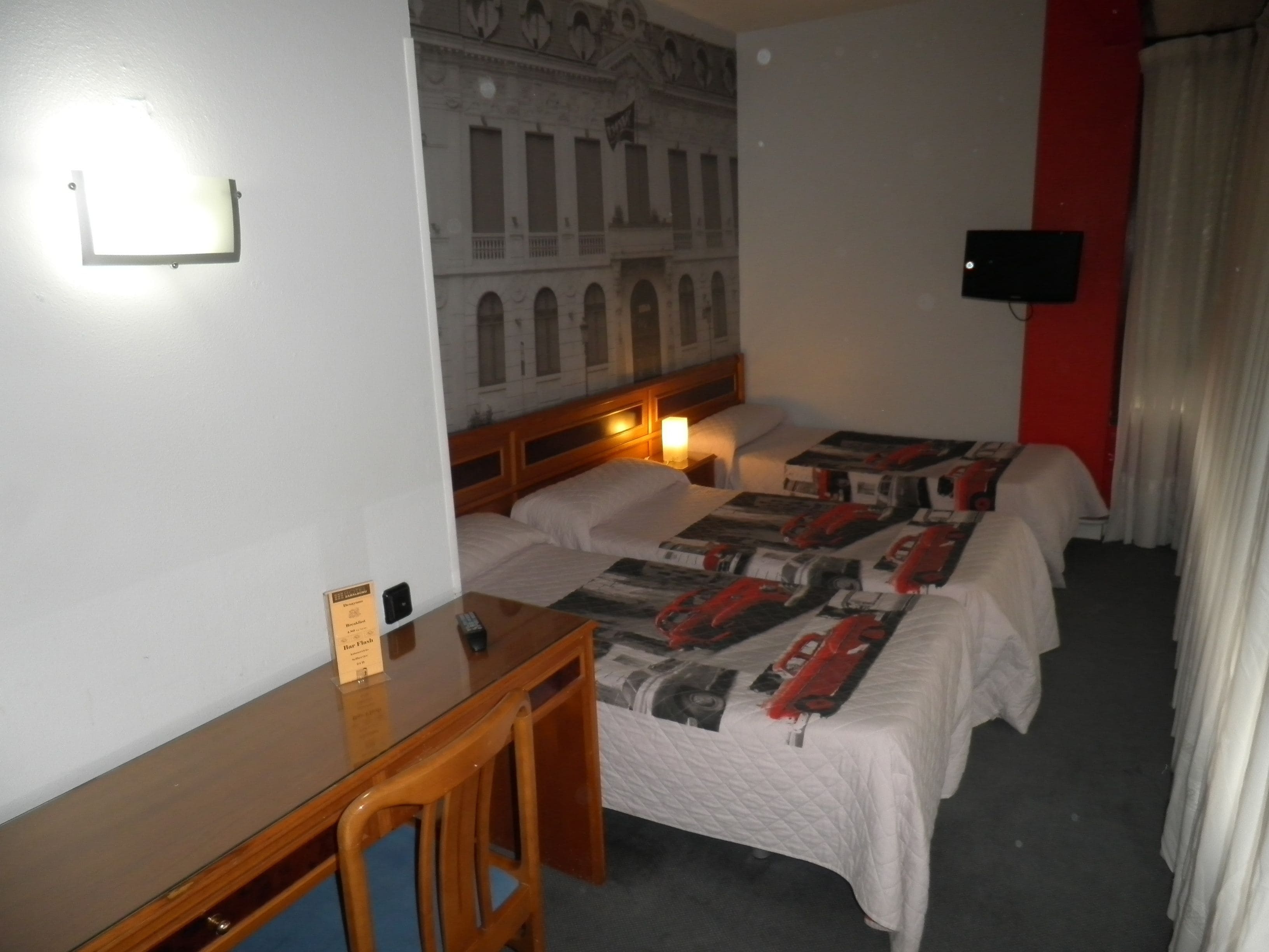 Habitación de hotel con dos camas, un escritorio, una silla, un televisor de pantalla plana y una lámpara de pared.